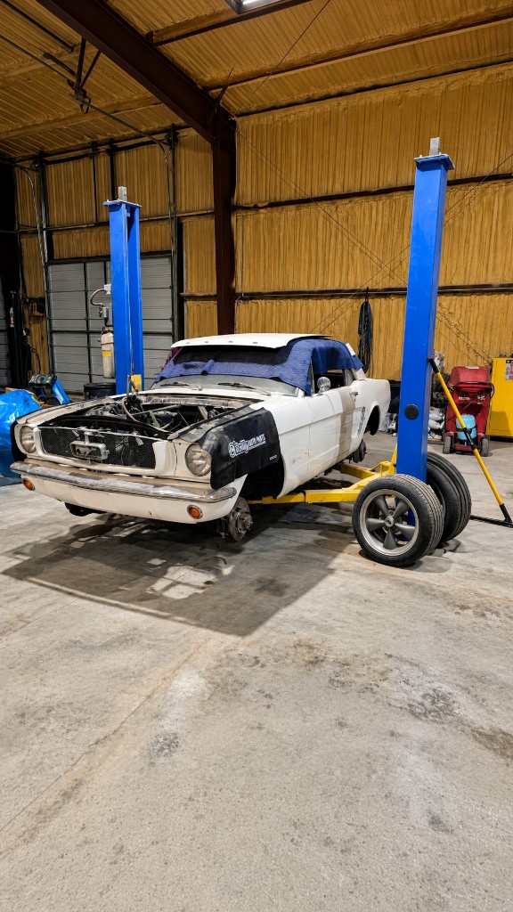 White classic Mustang with the front clip off, raised on a lift in a repair shop
