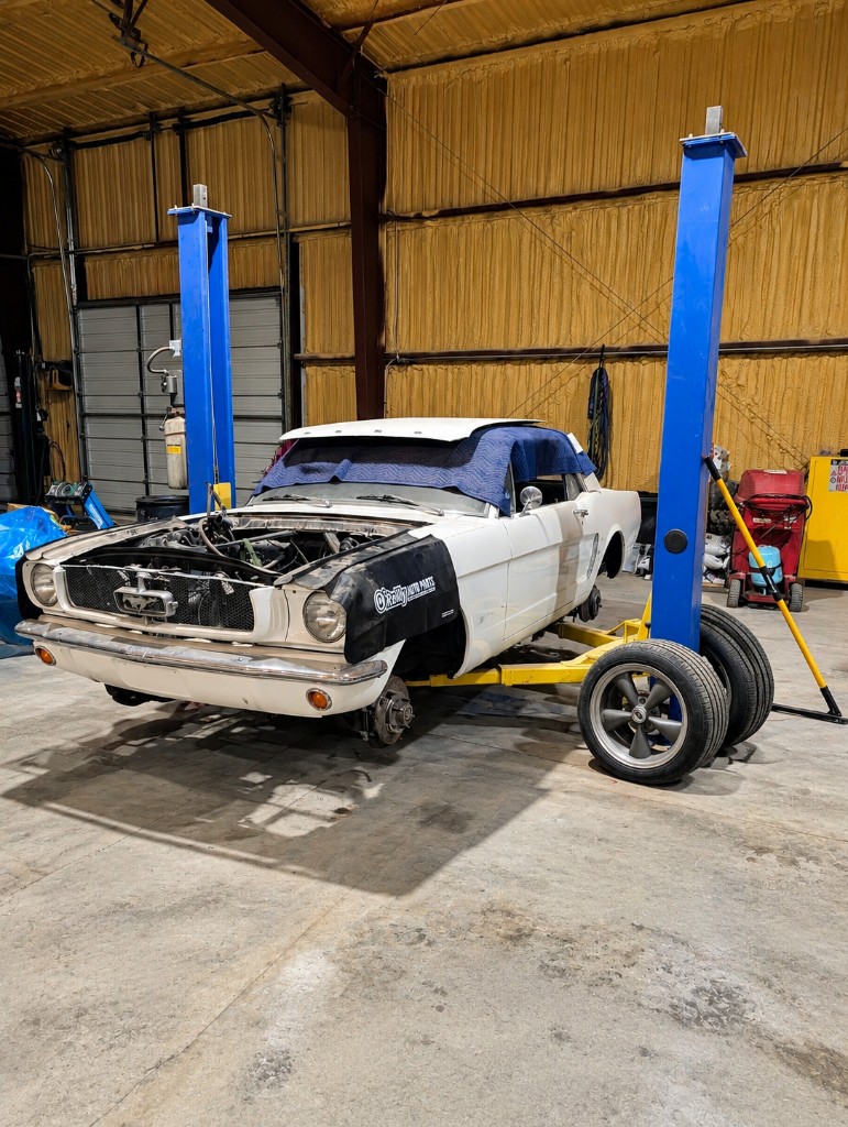 White classic Mustang with the front clip off, raised on a lift in a repair shop
