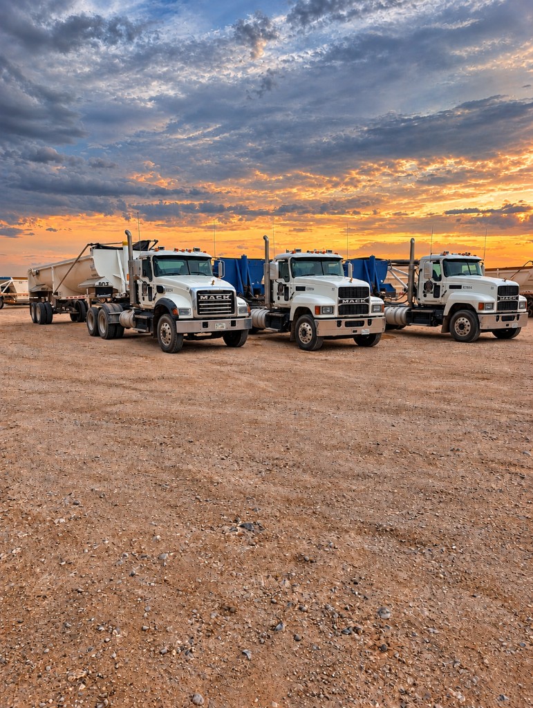 White Mack trucks parked on a gravel yard at sunset, one attached to a light-colored trailer